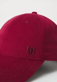 Căciulă roșie din material textil, cu o margine ușor curbată. Prezintă o suprafață texturată și un mic logo embosat în formă rectangulară.