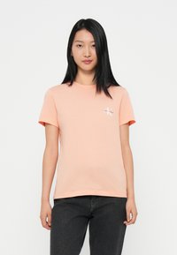 Light pink cotton t-shirt with a crew neck, short sleeves, and a white Calvin Klein logo on the left chest. Paired with dark jeans.