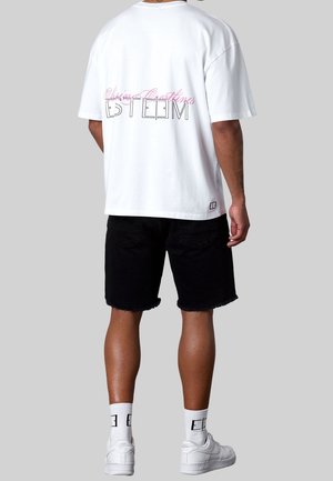 Homme portant un t-shirt blanc oversize avec le texte "Esteem", un short noir, des chaussettes blanches et des baskets blanches, debout devant un fond gris.