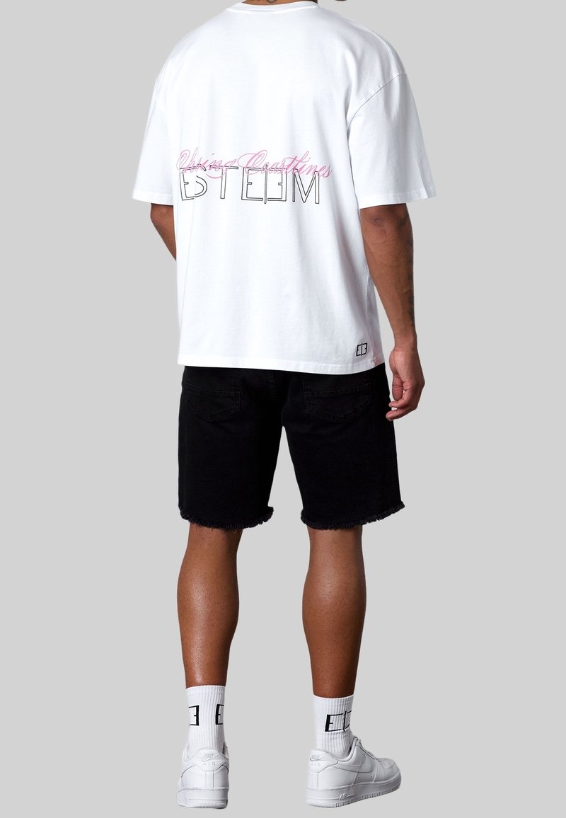 Homme portant un t-shirt blanc oversize avec le texte "Esteem", un short noir, des chaussettes blanches et des baskets blanches, debout devant un fond gris.