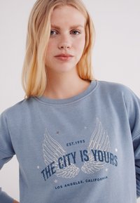 Felpa azzurra chiara con maniche lunghe. Il design presenta ali stampate e testo in blu scuro, con accenti di stelle argentate. Texture morbida.