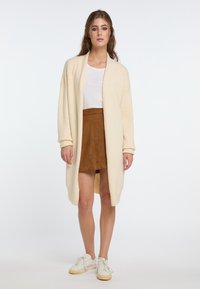 Kreminė atvira kardiganas, pagamintas iš minkštos medžiagos su ilgomis rankovėmis, derinamas su rudomis zomšos mini sijonu ir baltais sneakersais.