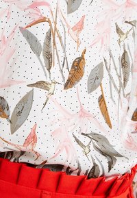 Top bianco con un motivo floreale e di uccelli in rosa, marrone e grigio, su uno sfondo nero a pois, abbinato a una cintura rossa texturizzata.