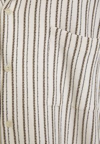 Randig skjorta med knappar i vitt med tunna bruna och svarta ränder; har en bröstficka och texturerat tyg. Knapparna är ljusbeige.