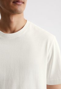 T-shirt en coton blanc avec un col ras du cou. Le tissu semble doux et lisse, avec des manches courtes et une coupe décontractée.