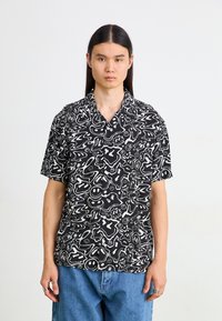 Camicetta nera a maniche corte con motivi bianchi astratti, dotata di colletto a punte e bottoni, abbinata a pantaloni in denim blu.