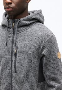 Άνδρας που φοράει γκρι φούτερ με φερμουάρ και υφή, με κουκούλα με κορδόνι και καφέ κυκλικό έμβλημα στο αριστερό μανίκι, μπροστά σε λευκό φόντο.