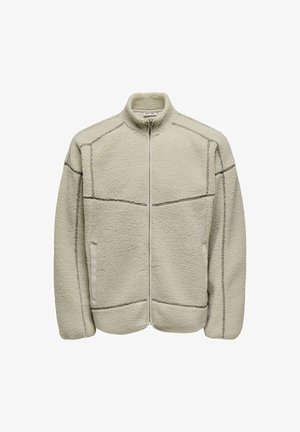 Polarowa kurtka w jasnym beżu z wysokim kołnierzem, z zamkiem błyskawicznym z przodu i dwiema bocznymi kieszeniami. Charakteryzuje się kontrastowymi ciemnymi szwami wzdłuż szwów.