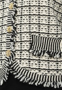 Černo-bílý pletený cardigan s texturovaným vzorem, zlatými knoflíky, třásněmi na okrajích a proužkovanou kapsou.