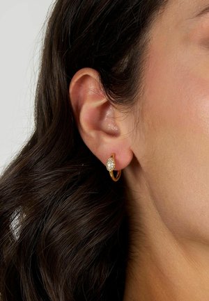 Orecchini a cerchio in oro con un charm ovale testurizzato e finitura scintillante, sullo sfondo di capelli e pelle scuri.
