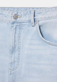 Lys blå denimjeans close-up, der viser linning, metalknap, bæltestrop og forlomme med synlige syningsdetaljer.