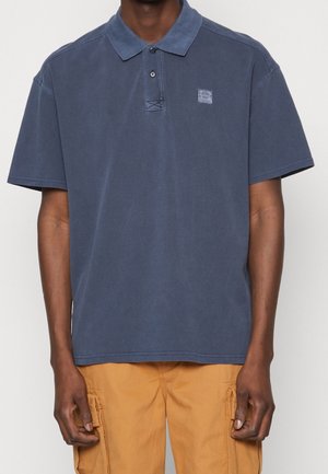 Mężczyzna w wyblakłej, granatowej koszulce polo z krótkim rękawem, zapinanej na dwa guziki, oraz w musztardowych spodniach typu cargo na jednolitym tle.