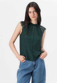 La modelo lleva un top sin mangas de color verde oscuro con encaje, combinado con jeans azules de cintura alta, posando con confianza con las manos en los bolsillos.