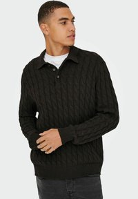 Donkerbruine gebreide trui met een verhoogd kabelpatroon, knoopsluiting bij de kraag en geribbelde manchetten en onderrand. Draagt over een wit t-shirt.