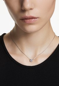 Sudraba ķēdītes kaklarota ar mazu, apaļu piekariņu, kas rotāts ar mirdzošu akmeni, valkāta pret melnu ribotu krekliņu.