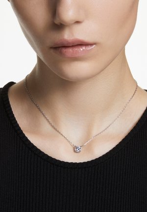 Zilveren ketting met een kleine, ronde hanger met een schitterende steen, gedragen tegen een zwart geribd shirt.