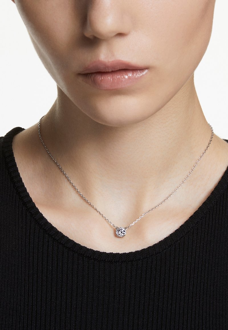 Sudraba ķēdītes kaklarota ar mazu, apaļu piekariņu, kas rotāts ar mirdzošu akmeni, valkāta pret melnu ribotu krekliņu.