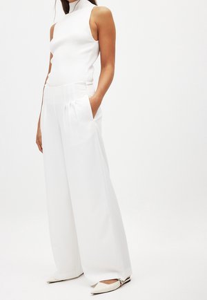Femme portant un haut blanc sans manches à col montant, un pantalon blanc à jambes larges plissées, et des ballerines blanches pointues avec des brides à la cheville.