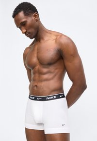 Αθλητικός άνδρας με μυώδη σωματότυπο που φοράει λευκά αντρικά εσώρουχα Nike με μαύρη ζώνη μέσης με λογότυπο, στέκεται με το κεφάλι σκυφτό.