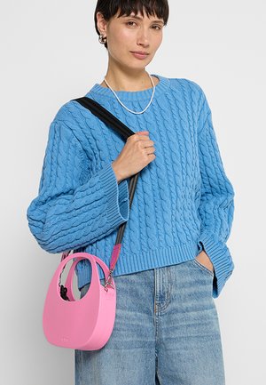 Borsa a mano rosa con forma arrotondata, realizzata in materiale liscio. Presenta una tracolla nera e dettagli decorativi, indossata da una persona in un maglione lavorato a maglia blu.