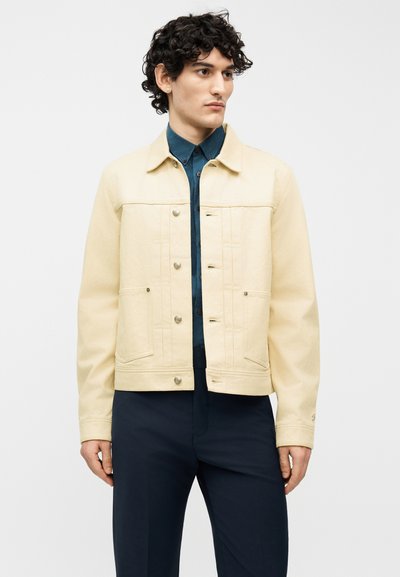 Νέος άνδρας με σγουρά μαλλιά, φοράει μια κρεμ ζακέτα με κουμπιά πάνω από ένα σκούρο μπλε πουκάμισο και ναυτικό μπλε παντελόνι, στέκεται μπροστά σε ένα απλό φωτεινό φόντο.