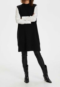 Abito nero senza maniche con collo alto, indossato sopra una camicia bianca a maniche lunghe. Completato con pantaloni neri aderenti e stivaletti con tacco a punta.