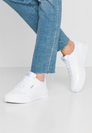 Person iført blå jeans og hvide Puma sneakers, stående med den ene fod let hævet på en lysegrå gulv.