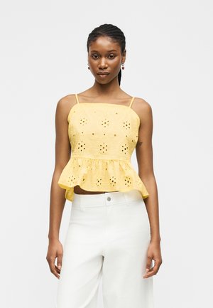 Femme portant un haut jaune sans manches en œillet avec un ourlet à volants et un pantalon blanc taille haute, debout devant un fond uni.