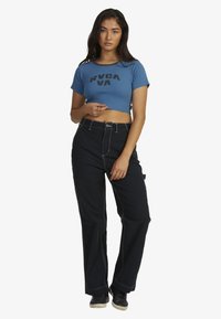 Blå cropped T-shirt med sort "RVCA" logo, parret med sorte bukser med hvid syning og sidelommer. Sorte sko afslutter outfittet.