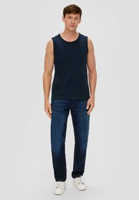 Marineblaue ärmellose Shirt aus Baumwollmischung, kombiniert mit dunkelblauen Jeans. Das Model steht vor einem hellgrauen Hintergrund und trägt weiße Sneakers.