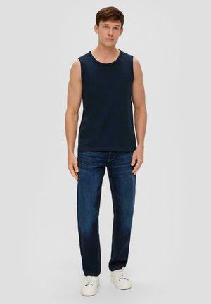 Marineblaue ärmellose Shirt aus Baumwollmischung, kombiniert mit dunkelblauen Jeans. Das Model steht vor einem hellgrauen Hintergrund und trägt weiße Sneakers.
