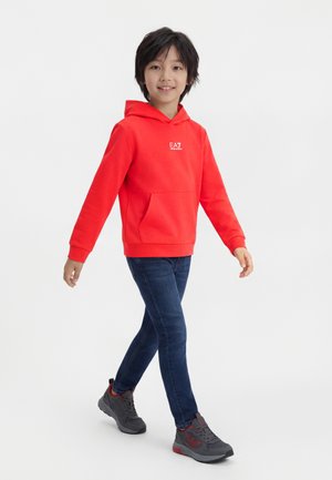 Lapsi kävelee päällään kirkkaan punainen hupullinen collegepaita, tummansiniset farkut ja harmaat tennarit valkoista taustaa vasten.