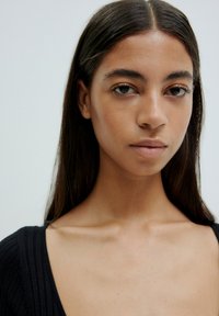 Nahaufnahme einer Person mit langen, glatten, dunklen Haaren und einem neutralen Ausdruck, die ein schwarzes, geripptes Top mit tiefem Ausschnitt trägt.