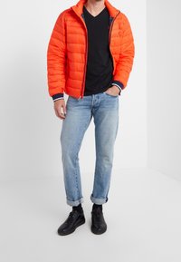 Muž v jasně oranžové péřové bundě přes černé triko s výstřihem do V, světlých modrých zkrácených džínách, černých ponožkách a černých teniskách stojící s rukou v kapse.