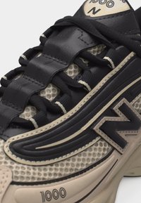 Κοντινό πλάνο αθλητικού παπουτσιού με υφή σε μαύρο και μπεζ χρώμα, με μαύρα κορδόνια, λογότυπο με το γράμμα «N» και εκτύπωση «1000» στη μπεζ σόλα.