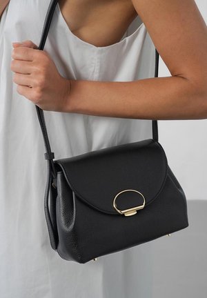 Femme en robe blanche tenant un sac à bandoulière en cuir noir avec une boucle ovale dorée et une sangle réglable.