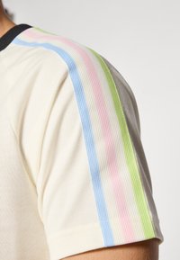 Chemise beige clair avec des rayures horizontales en bleu, rose et vert sur les manches ; fabriquée en tissu texturé avec un col rond noir.