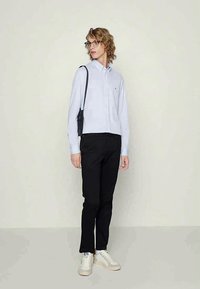 Ανοιχτό μπλε πουκάμισο με μικρό λογότυπο, μαύρο παντελόνι και λευκά αθλητικά παπούτσια. Η φιγούρα κρατά μια μικρή μαύρη τσάντα crossbody.