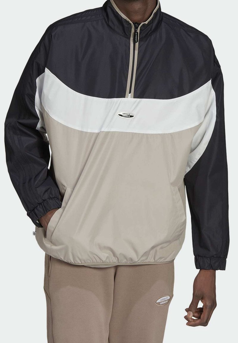 Man med en färgblockerad pulloverjacka med dragkedja i kragen och beige byxor, hand i fickan på jackan, varumärkeslogotyp synlig på bröstet och byxorna.