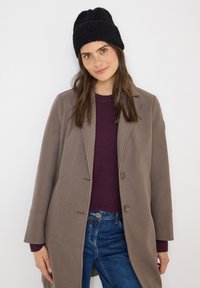 Harmaa takki, jossa on suuret kaulukset, kaksi nappia ja sivutaskut, käytetään tumman purppuran värisen ribbipaidan päällä. Musta neulottu pipo päässä.