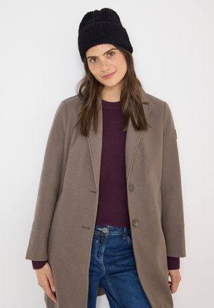 Grå frakke med store revers, to knapper og sidelommer, båret over en mørk lilla ribbet sweater. Sort strikket hue på hovedet.