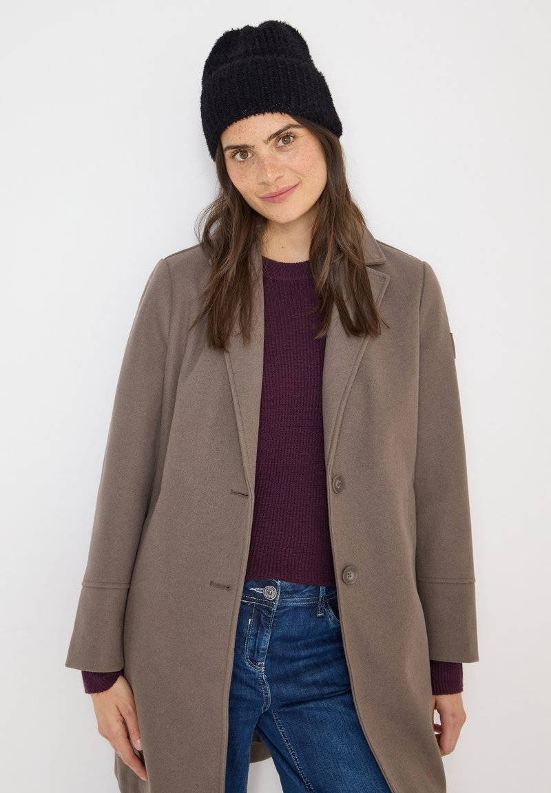 Harmaa takki, jossa on suuret kaulukset, kaksi nappia ja sivutaskut, käytetään tumman purppuran värisen ribbipaidan päällä. Musta neulottu pipo päässä.