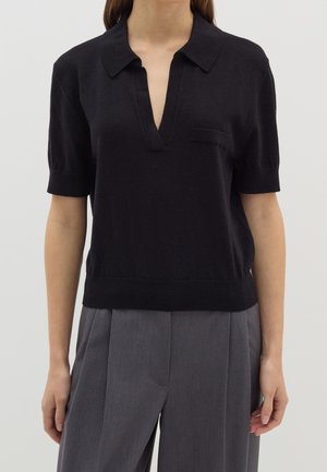 Sort kortærmet top med V-udskæring, tekstureret stof og en lille brystlomme. Båret med grå, plisserede bukser.