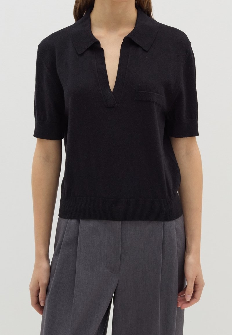 Haut noir à manches courtes avec un col en V, tissu texturé et petite poche poitrine. Porté avec un pantalon plissé gris.