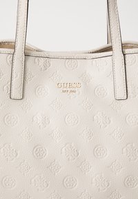 Sac fourre-tout beige texturé avec motif de logo en relief, présentant une plaque logo en or, des poignées doubles et un design structuré.