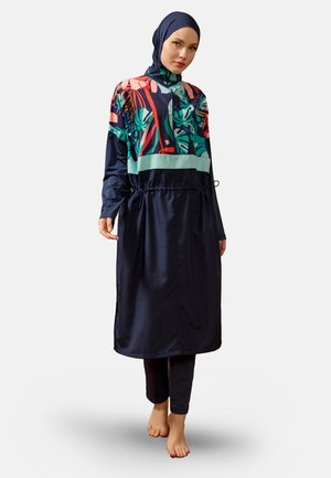 Donna con un vestito lungo blu navy con motivo floreale e hijab abbinato, in piedi a piedi nudi contro uno sfondo chiaro uniforme.