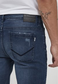 Jeans in denim blu scuro con taglio dritto. Presentano un'etichetta nera di marca, usura discreta e un design distintivo della tasca posteriore.