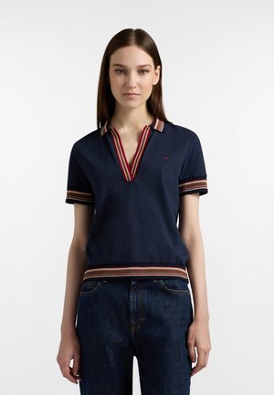 Donna che indossa una polo a maniche corte blu navy con colletto, maniche e orlo a righe rosse, bianche e beige, abbinata a jeans blu scuro.