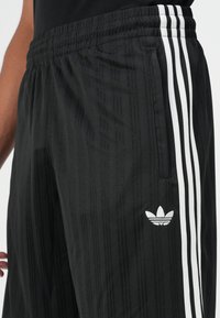 Black athletic pants with vertical striped texture, featuring white stripes on the sides and an embroidered logo on the lower leg.