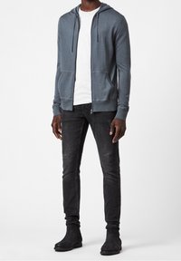 Sweat à capuche zippé gris avec des poches avant, porté sur une chemise blanche. Associé à un jean noir et des bottines noires. Design décontracté et confortable.